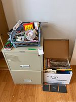 Filing cabinet with tote of office supplies on top, boxes of books beside