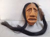 Full view of carved wooden mask with long hair strand displayed on white background