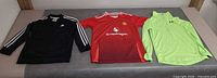 Three youth clothing items laid flat: Adidas black track jacket, red Manchester United jersey, neon green Under Armour pullover