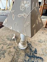 Full view of small lamp showing white base, cord, and embroidered taupe shade