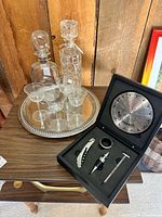All items arranged on wooden table: tray with glasses, two decanters, bar compass tool set open in box