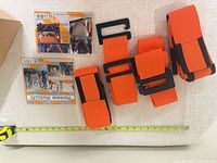 Pair of orange Forearm Forklift straps with black buckle loops, folded, next to packaging and measuring tape