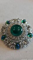 Full view of brooch showing central green cabochon, clear crystal halo and outer green-blue cabochons