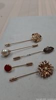 Five of the six gold-tone stick pins laid out showing overall design