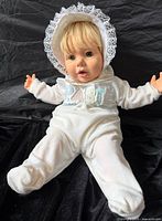 Doll seated wearing white hooded onesie with BABY applique and lace bonnet