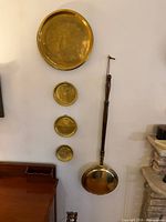Wall-mounted display showing four brass trays in descending size plus brass bed warmer with wood handle