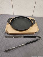 Round Weber cast iron griddle atop cardboard with tongs in foreground