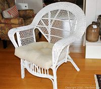 White wicker armchair with beige cushion sitting on hardwood floor. Angle shows curved backrest and armrests with lattice weave.