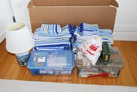 Full lot photo showing the folded towels, table lamp on the left, first aid kit (blue box labeled 'First Aid'), and clear sewing box with supplies inside, all placed in front of a cardboard box on wooden floor.