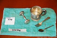 Photo showing sterling silver baby cup, baby fork, baby spoon, and baby rattle on turquoise cloth marked 'Sterling' with MaxSold lot tag.