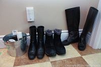 Three pairs of black ladies boots in a row and a metal bucket with boot care items on floor.