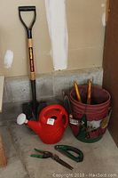 Photo showing garden shovel, red watering can, gloves, pruning shears, and a red bucket with apron containing multiple garden hand tools.
