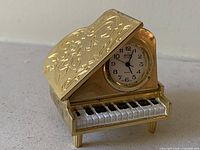 Front view of gold-tone piano clock with lid partially open showing dial and keyboard