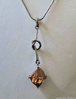 Close view of pendant showing square princess-cut orange-brown topaz in four-prong silver-tone setting, knotted bar detail, portion of snake chain