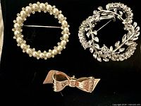 Front view of three brooches: faux-pearl wreath, leaf rhinestone wreath, gold-tone bow