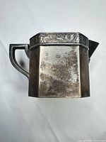 Side view of tarnished octagonal metal creamer showing decorative Egyptian motif band, handle and spout