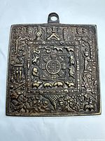 Front of square brass plaque showing detailed Tibetan motifs and hanging loop