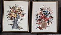 Two of the three embroidered pieces side by side showing floral pitcher and floral basket designs within gold-tone frames
