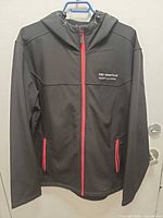Front view of black softshell jacket showing red zipper, pocket zippers and embroidered logo
