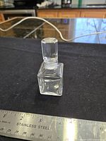 Full view of square glass perfume bottle with stopper next to ruler