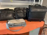 Group shot of three bags: gray messenger front, two black bags behind including rolling suitcase