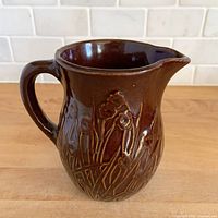 Full view of brown glazed stoneware pitcher showing embossed floral detail, handle, and spout