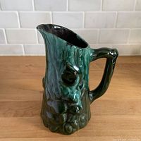 Left-front view of green drip glazed ceramic pitcher showing mushroom relief design and spout