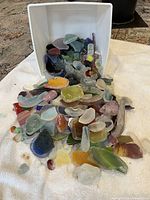 Bulk pile of multi-coloured tumbled glass spilling from white bin onto towel