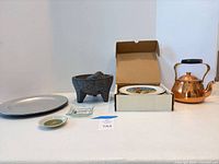 Entire lot displayed on table: charger, small dish, packaged item, molcajete, boxed plate, copper kettle