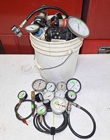Bucket filled with assorted pressure gauges, regulators and hoses; additional gauges arranged on table in foreground