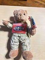 Front view of tan plush teddy bear in desert camo uniform, Pep Boys gift card tag on chest, holding American flag