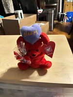 Front view of red plush teddy bear holding purple heart with tag visible