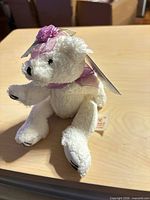Front left view of small white plush bear with pink ribbon and flower, hang tag visible