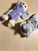 Beige teddy bear with purple scarf and gray hippo plush on wooden surface