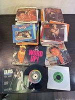 Top-down view of stacked 7" singles showing titles such as Aretha & Whitney, Billy Idol, Cameo, Elvis and Grease