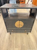 Front view showing black wood cabinet with open shelf, twin doors, circular brass medallion, lower ring pulls
