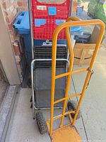Overall view of orange hand truck ahead of stacked folding hand truck and step stool
