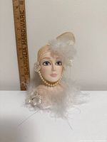 Front view of porcelain flapper bust showing face, pearls, feathers and headpiece with ruler for scale
