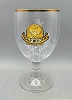 Close-up of a Grimbergen beer glass showing gold rim, phoenix logo, goblet shape, footed stem