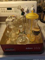 Overall view of box containing assorted glass jars, bottles, decanters, yellow-lid jar and wine charms