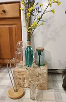 Group shot showing teal bottle with flowers, clear stopper jar, aqua jar with cork/spoon, square vase with lunaria, wood/chrome hook stand