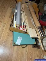 Cardboard box atop green case holding dowels, wood sheets, plastic film rolls