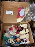 Open box showing multiple small dolls, three ceramic baby shoes, and bag of accessories