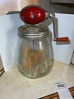 Full view of glass butter churn showing jar, metal lid, red crank housing, wooden paddles