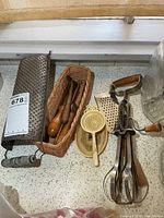 Overview of all utensils including box grater, wicker basket with wood-handled tools, plastic strainer set, egg beater