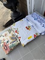 Three folded textile pieces laid out: floral fabric panel on left, yellow/red floral-fruit tablecloth centre, blue/white tablecloth right