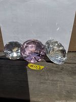 Three faceted glass diamond-shaped paperweights, two clear and one purple, placed side by side