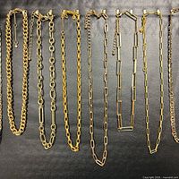 Nine gold-tone chain necklaces laid side by side on black background showing various link types