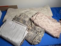 Overview of four fabric pieces laid out: woven cream roll, geometric print, paisley jacquard, silvery satin