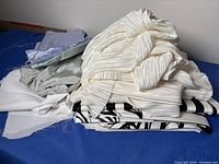 Stack of assorted fabrics showing ivory pleated, mint satin, lavender, plain white, and black-patterned pieces